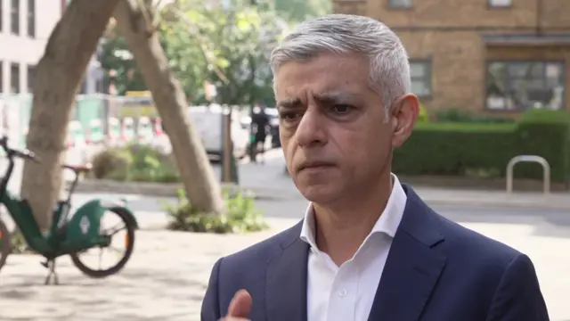 Sir Sadiq Khan stands outside on a sunny day being interviewed. He is wearing a dark suit and a white shirt.