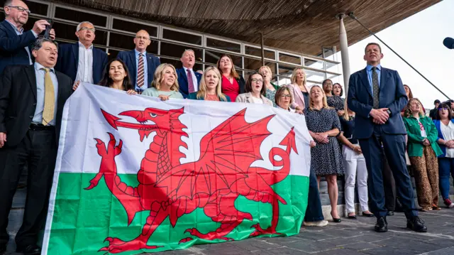 Rhun is standing next to a rows of people, some of whom are holding up a large Wales flag