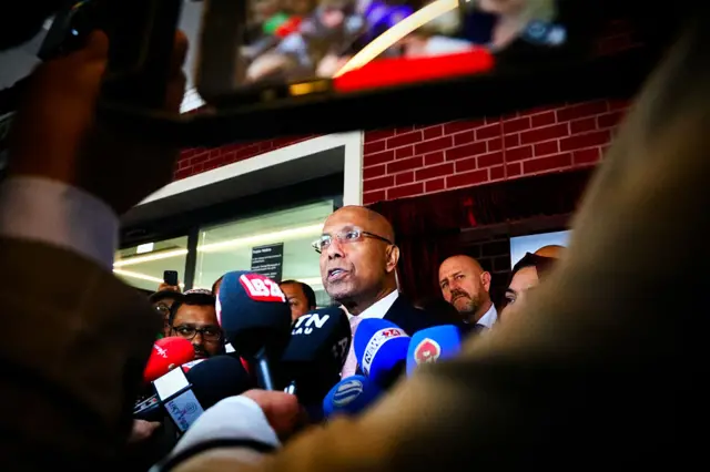 A politician in a dark suit speaking in front of a bank of microphones