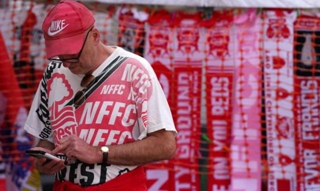 A man wearing a Nottingham Forest t-shirt and hat on his phone