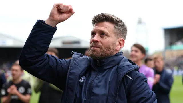 Luke Garrard wearing a blue overcoat raises his right fist in celebration after Boreham Wood have beaten Carlisle in the play-offs