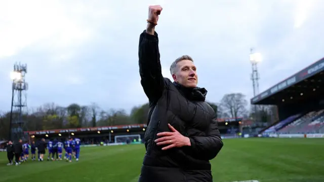 Rochdale boss Jimmy McNulty wearing a black overcoat punches the air with his right fist after his side have beaten Wealdstone