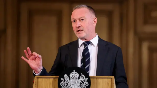 Headshot of Reed who speaks at a government lectern and holding one hand up gesturing