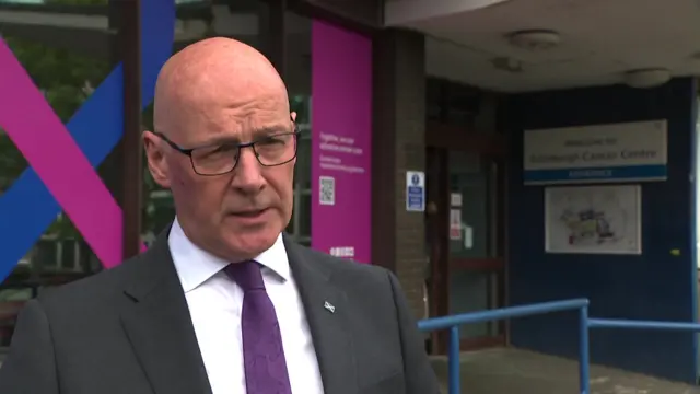 Scottish first minister John Swinney answering questions in a black suit, white shirt and purple tie outside a hospital