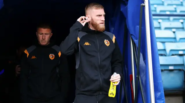 Oli McBurnie holds his hand to his ear while walking out of the tunnel to inspect the pitch at The Den before the game