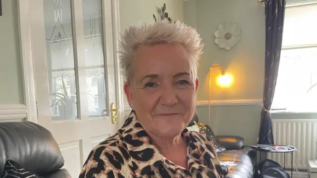 Lorraine Beavers wearing a leopard print blouse as she smiles and sits on a sofa in a living room in front of a door, window and lamp