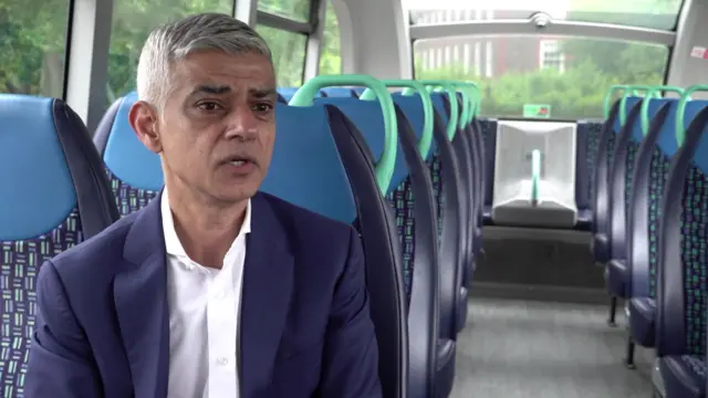 Sadiq Khan is wearing a navy blue jacket and a white shirt. He is sat down on a bus.