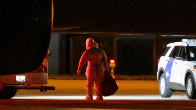 someone walking in a biohazard suit carrying a large bag underneath a plane with a police car beside them