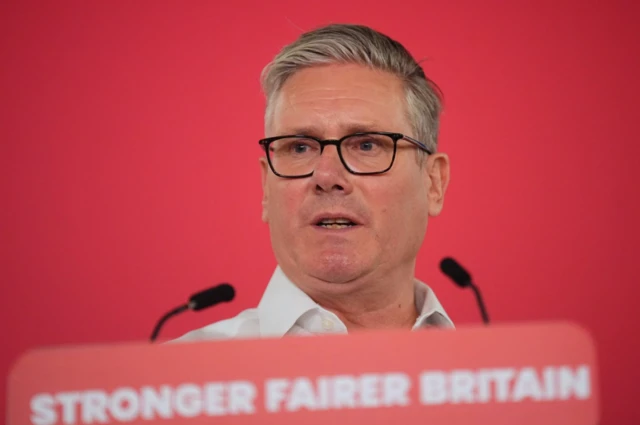 Starmer in a white shirt mid-speech in front of a lectern with two thin black mics on top and a sign reading "Stronger fairer Britain" in white on red background. There's a red wall behind him