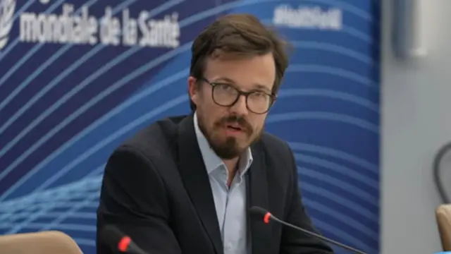 Dr Olivier le Polain sits behind a microphone, wearing a dark suit jacket and light-coloured shirt.