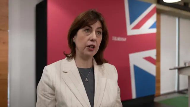 Lucy Powell in striped white blazer and grey top speaking in a room, a panel painted with a partial Union Jack flag behind her