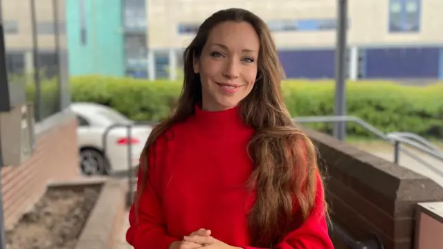 Catherine Atkinson, a white woman with long brown hair in a red jumper smiling at the camera.