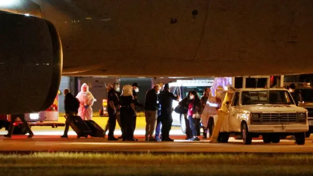 people standing around, including one in a biohazard suit, underneath an airplane