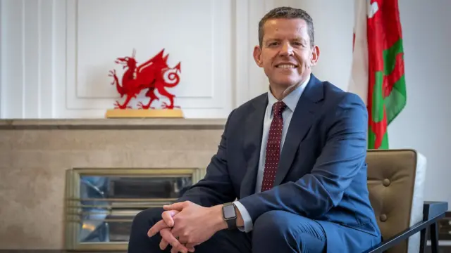 Rhun ap Iorwerth sits on a chair with a Welsh flag and a red dragon statue behind him. He wears a blue suit and smiles at the camera
