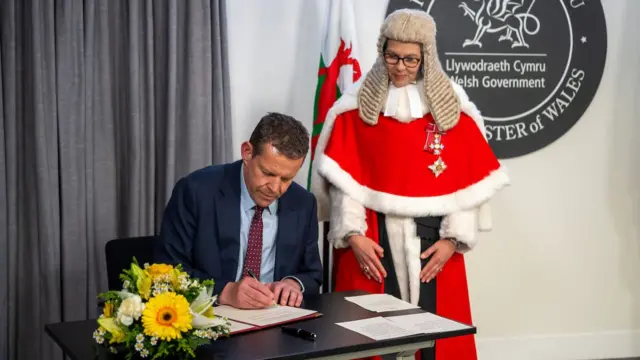 Rhun ap Iorwerth signs a document at a desk as a High Court judge stands watching over him.