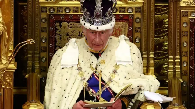 King Charles is seated on a throne wearing a large crown and robes. He is reading aloud from a booklet in his hands.