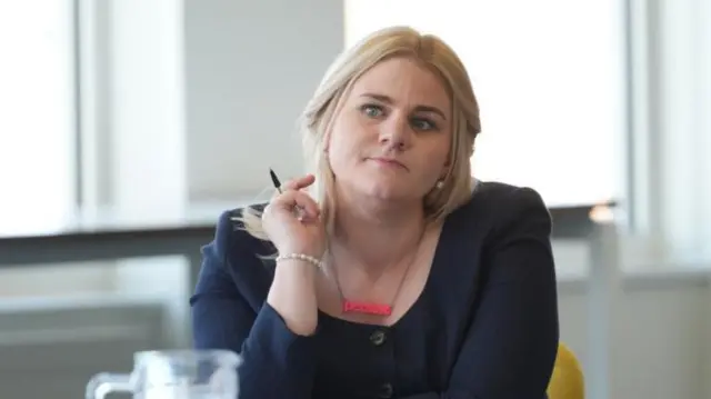 Alex Davies-Jones is pictured sitting at a table wearing a dark blue outfit, and a necklace with pink lettering that says 'persist'. She is holding a pen in her right hand and looking off to one side.