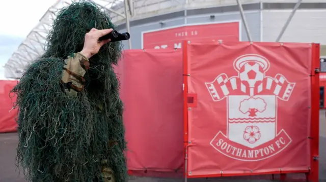 A man in army fatigues and camoflage gear holds a pair of binoculars outside of St Mary's