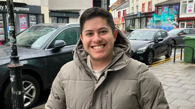 Sam Sejovia stands on a street in Bangor wearing a grey coat. He has short black hair.