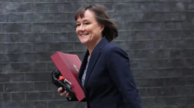 Jo Stevens walks outside carrying a red folder. She is wearing a navy suit and has a short brown bob.