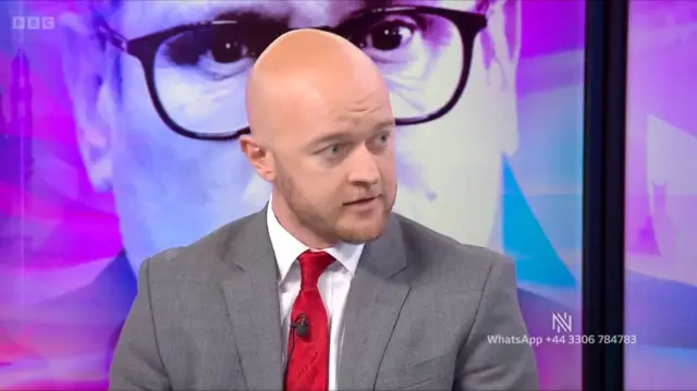 Jonathan Hinder wearing a ruby red tie and grey suit jacket speaking on Newsnight