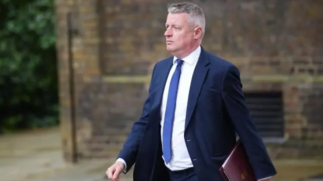 Luke Pollard is shown walking outdoors wearing a suit and tie, with a dark red folder under his left arm