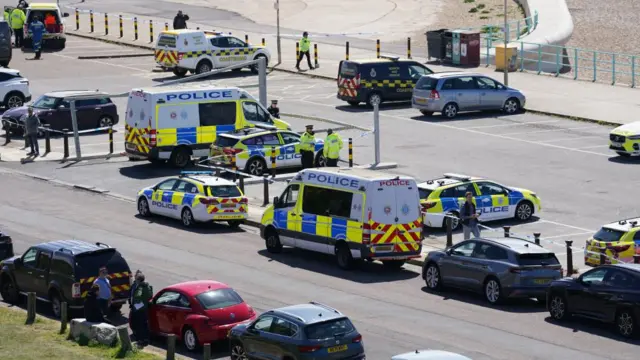 A large gathering of emergency vehicles, including police vans, police cars and coastguard vans, can be seen in a car park. Police tape can be seen stretching across multiple bollards.