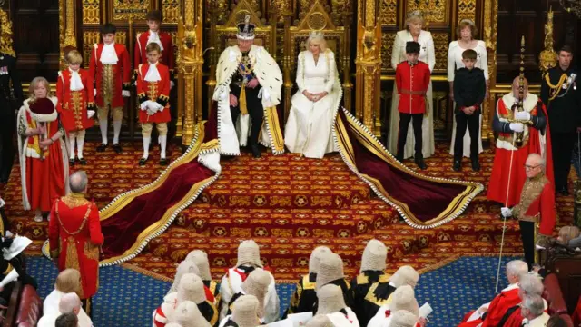 King Charles III, wearing the Imperial State Crown and the Robe of State, alongside Queen Camilla, wearing the George IV State Diadem and an embroidered gown by Fiona Clare, in the Chamber of the House of Lords during the State Opening of Parliament