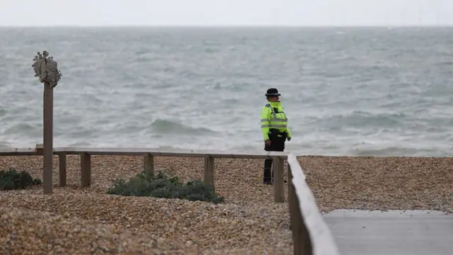 A police officer seen standing on a beach, wearing a luminous jacket. The sky is grey and waves appear choppy