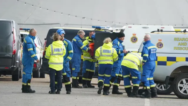Multiple members of emergency services, dressed in dark blue and luminous jackets, gather around emergency vehicles in a car park.