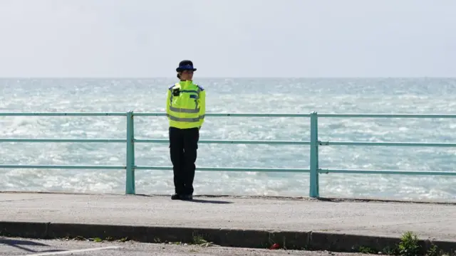 A police officer stands at a pier while in uniform, wearing a luminous jacket and dark cap. The sea can be seen behind her.