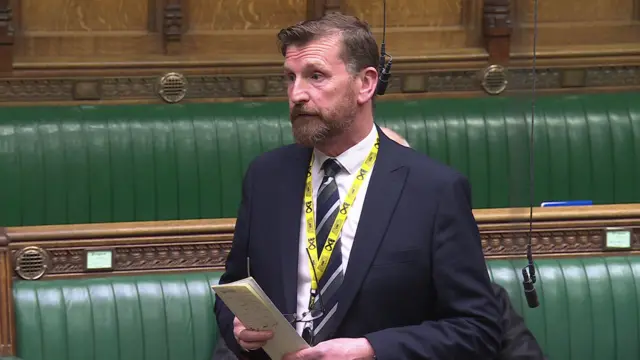 Dave Doogan stands in the House of Commons, wearing a dark suit and dark striped tie. He has a yellow lanyard around his neck