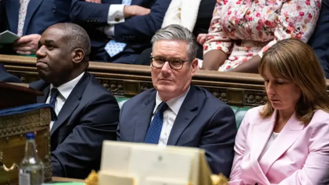 Keir Starmer sits between David Lammy and Rachel Reeves in the House of Commons. He is wearing a dark suit and navy tie.
