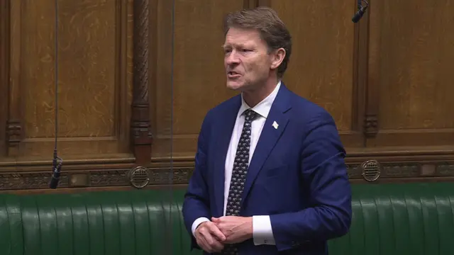 Richard Tice wears a dark blue suit while speaking in the House of Commons. He is wearing a dark patterned tie.