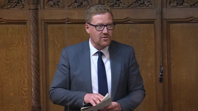 Jonathan Brash stands in the House of Commons, wearing a grey-blue suit and a navy tie. He is holding a set of papers in his hand