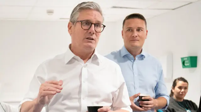 Keir Starmer in a white shirt at the forefront of the frame, with former health sec Wes Streeting looking at him from behind. They're in a white-walled hospital room