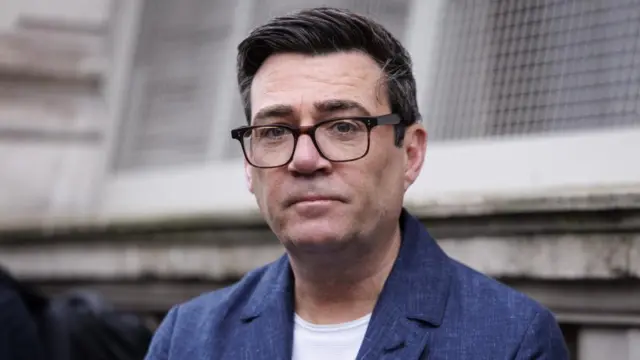 Andy Burnham pictured in March, he is wearing a blue jacket and square glasses