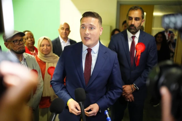 Wes Streeting in a suit surrounded by voters at the election count in Redbridge. He's wearing a dark blue suit, white shirt and burgundy tie