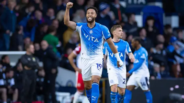 Kyle Woottonof Stockport County celebrates after scoring a goal wearing a blue Stockport shirt with his right arm raised and fist clenched