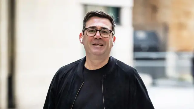 Andy Burnham walks down a street wearing a black t-shirt and black jacket.