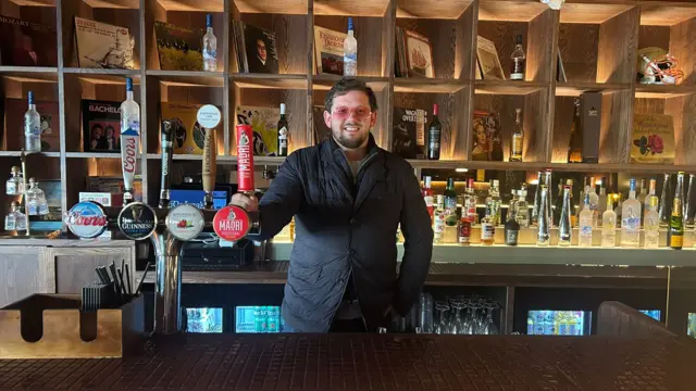A man in rose-pink shades and a black jacket stands smiling behind a bar.