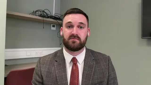 Matthew Torbitt sits in a plaid suit jacket and red tie. He is wearing AirPods and is sitting in a room with a light green wallpaper.