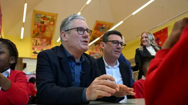 Keir Starmer and Andy Burham sitting at a primary school table with some pupils in red jumpers