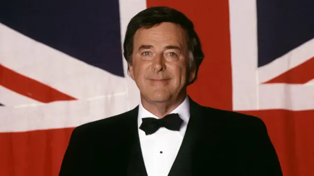 Terry Wogan in a suit and bow tie in front of a Union flag