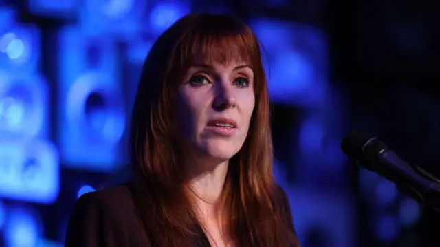Close up of former deputy PM Angela Rayner on stage, a mic in front of her, a blue screen in the blurred background
