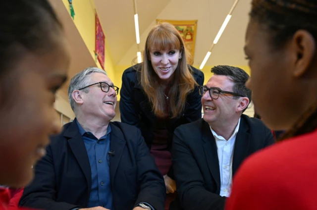 Keir Starmer sitting down (L) looking up smiling to Angela Rayner (C), who is leaning over the PM and a laughing Andy Burnham (R). In the foreground, framing the trio, are two pupils in red sweaters