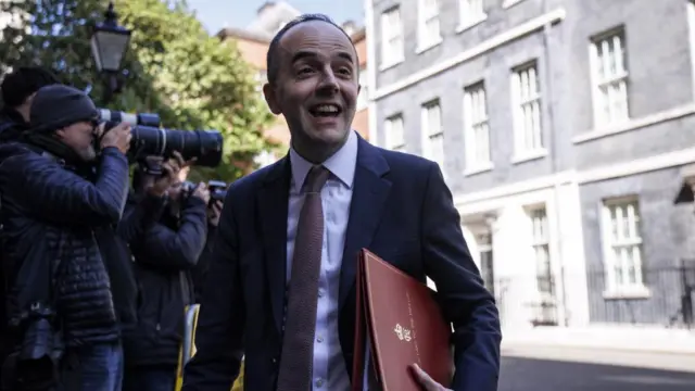 James Murray walks outside Downing Street with several photographers visible behind him. He is wearing a navy suit and has a red binder under his arm.
