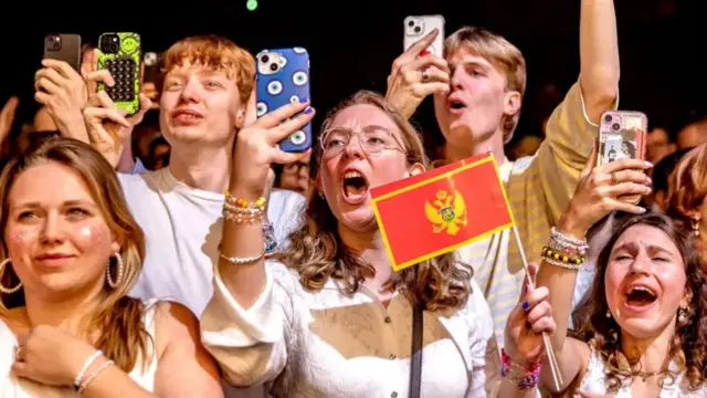 A group of people in a crowd looking towards the camera and cheering, some are holding phones up to record.