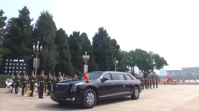 A black car with US and China flags drives past a military band