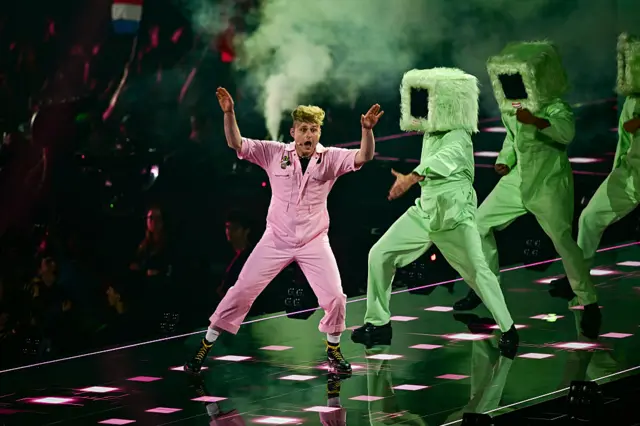 Look Mum No Computer on stage at Eurovision in pink, three people in green with furry boxes on head behind him.